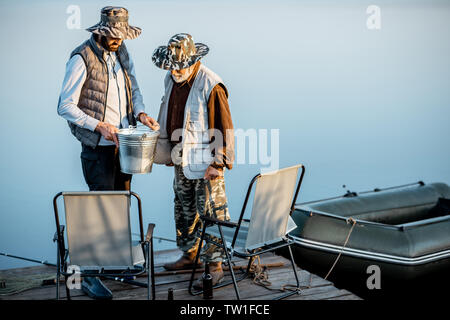 Gerne Großvater mit erwachsenen Sonne stehen zusammen mit frisch gefangenen Fisch auf der Pier in der Nähe des Sees in den frühen Morgenstunden Stockfoto