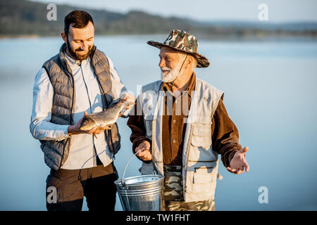 Gerne Großvater mit erwachsenen Sonne stehen zusammen mit frisch gefangenen Fisch auf der Pier in der Nähe des Sees in den frühen Morgenstunden Stockfoto