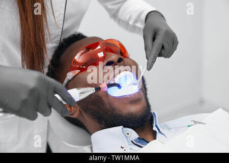 Afrikanischer Mann liegen auf zahnarztstuhl mit offenen Mund, Schutzbrille, während die weiblichen Zahnarzt tun Füllen mit UV-Licht. Mann Besuch Arzt heilen Zähne in der Klinik. Konzept der Stomatologie. Stockfoto