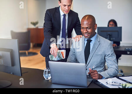 Zwei lächelnde diverse Geschäftsleute zusammen, die an einem Notebook arbeitet an einem Schreibtisch in einem hellen modernen Büro Stockfoto
