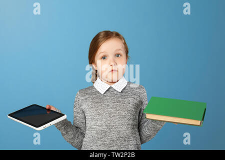 Fröhliche kleine Mädchen auf einem blauen Hintergrund. Das Kind hält ein Buch und eine Tablette. Der Begriff der Bildung, Schule, Auswahl, Vergleich. Stockfoto