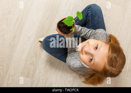 Blick von oben auf ein Kind sitzen auf dem Boden und Halten einer jungen Pflanze. Das Mädchen ist auf der Suche in die Kamera. Das Konzept der Ökologie, Natur, Pflege. Stockfoto