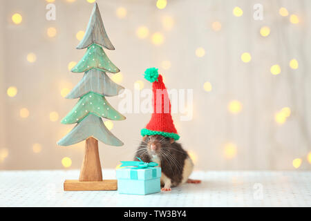 Süße Ratte in hat in der Nähe von dekorativen Weihnachtsbaum und kleines Geschenk auf Tisch gegen Defokussierten leuchtet Stockfoto