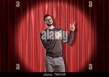 Crop Blick auf jungen gutaussehenden Mann in Freizeitkleidung im Rampenlicht stehend gegen roten Bühnenvorhang und nach oben mit beiden Händen. Stockfoto