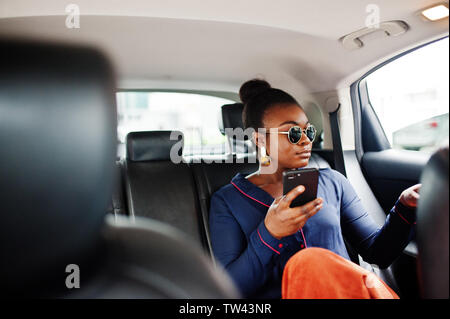 Reiche Geschäft afrikanische Frau mit Sonnenbrille sitzen an suv-Auto mit schwarzen Ledersitzen. Handy zur Hand. Stockfoto