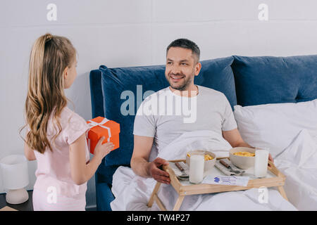 Entzückende Tochter holding Vatertag Geschenk, während in der Nähe von Pater Frühstück im Bett stehend Stockfoto
