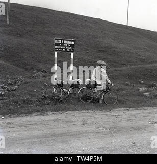 1950, historische, eine männliche touring Radfahrer stehen in seinem Regencape mit seinem Fahrrad neben einer Schotterpiste auf dem San Pellegrino (Passo S. Pellegrino), einen hohen Pass - 1918 Meter über dem Meeresspiegel - in den italienischen Dolomiten, ein Gebirgszug im nordöstlichen Italien. Stockfoto