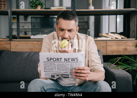 Schöner Mann essen frischen Apfel beim Lesen Sport Nachrichten Zeitung zu Hause Stockfoto