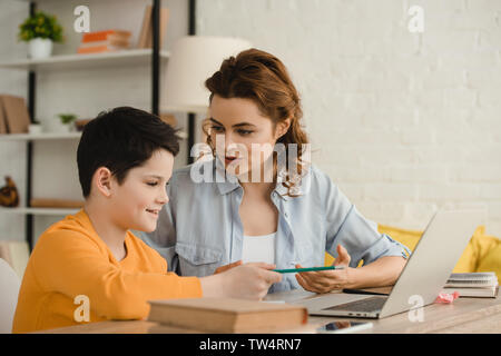 Lächelnde Mutter helfen adorable Sohn Hausaufgaben beim Sitzen am Schreibtisch mit Notebook Stockfoto