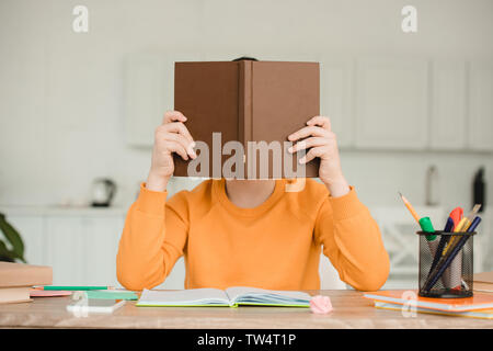 Junge versteckt sich hinter buchen Sie beim Sitzen am Schreibtisch und macht Hausaufgaben zu Hause Stockfoto