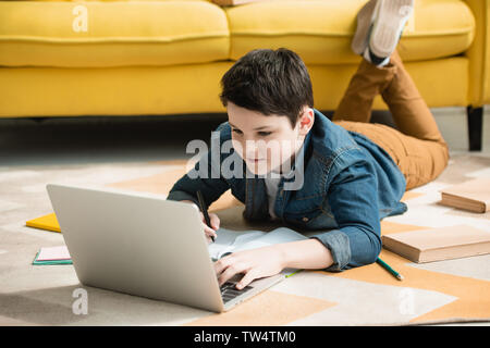 Aufmerksame Junge lag auf dem Boden in der Nähe der Bücher und mit Laptop während der Hausaufgaben Stockfoto