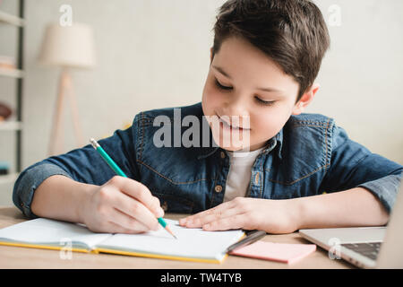 Fröhliche junge Schreiben im Notebook beim Sitzen am Schreibtisch und Hausaufgaben Stockfoto
