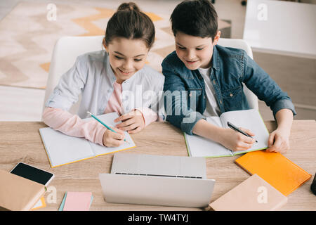 Lächelnd Bruder und Schwester schreiben in Notebooks und mit Laptop zusammen Stockfoto