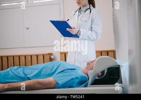 7/8-Ansicht von jungen Radiologen schreiben auf Zwischenablage beim Stehen in der Nähe von Patienten, der auf MRT-Scanner bed Stockfoto