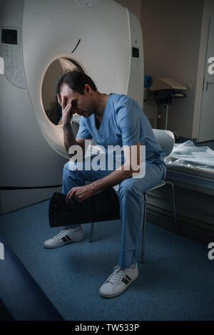 Erschöpft Radiologe holding Röntgendiagnostik, während in der Nähe des CT-Scanners im Krankenhaus sitzen Stockfoto