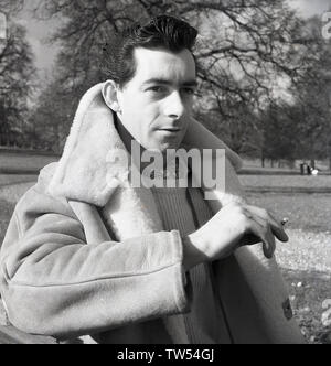 1960, historische, jung bin ein über die Stadt stehen in einem Park in den Moden des Tages, das Tragen eines crewneck Wollpullover, eine Krawatte, einen dicken sheepsking Jacke und rauchen eine Zigarette, England, UK. Stockfoto