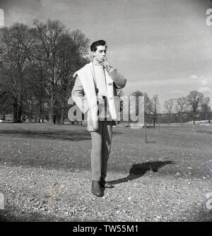 1960, historische, Fotograf... ein Junge bin ein über Stadt' mit seiner Filmkamera, draußen auf einem Schotterweg in einem Park in den Moden des Tages, das Tragen eines crewneck Wollpullover, eine Krawatte und einem dicken Schaffell Jacke und rauchen eine Zigarette, England, UK. Stockfoto