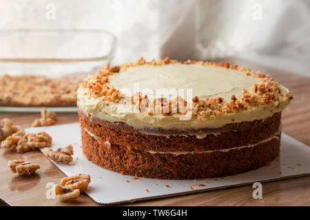 Die Karotte Kuchen mit Frischkäse Creme und geschreddert Walnusskerne gekrönt und ist auf einem Holztisch mit einer freien Version von Walnusskernen über den Tisch verstreut platziert Stockfoto