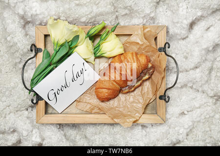 Fach mit leckeren Croissants, 'Guten Tag' Karte und Blumen auf dem Bett Stockfoto