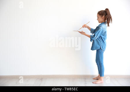 Kleines Mädchen mit Wasserfarben und Pinsel gegen weiße Wand im Innenbereich Stockfoto