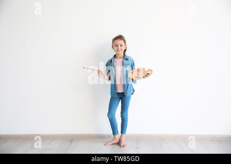 Kleines Mädchen mit Wasserfarben und Pinsel gegen weiße Wand im Innenbereich Stockfoto