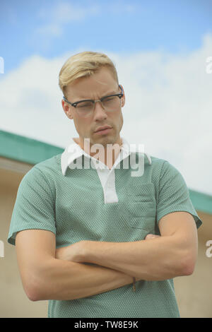 Ein weißer kaukasischer Mann, der ein grünes Poloshirt aus den 60er Jahren trägt und eine Brille aus den 60er Jahren trägt, die gekreuzt die Arme zeigt und denkend nach unten blickt. Stockfoto