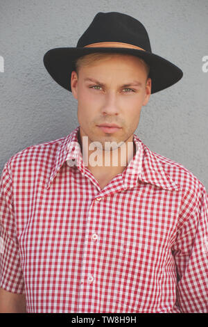 Ein blondes männliches Modell mit einem karierten Hemd in Western-Rot und einem Fedora-Hut, der sich an einer Wand im Freien aufsetzt. Ein Nahaufnahme-Headshot-Porträt. Stockfoto