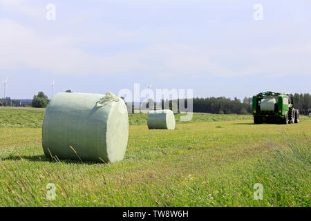 Silageballen in grün Kunststoff mit Traktor ziehen einer Ballenpresse Heu Feld an einem schönen Tag im Sommer. Fokus auf den Ballen auf der linken Seite. Stockfoto