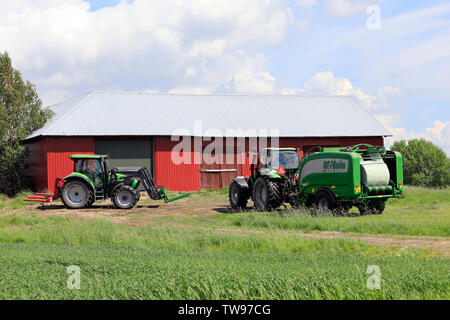 Salo, Finnland. Juni 15, 2019. Deutz-Fahr Traktoren und McHale Ballenpressen-Wickler-Kombination bei der Arbeit im Heu Feld vor der roten Scheune an einem Tag im Sommer. Stockfoto