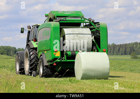 Salo, Finnland. Juni 15, 2019. Deutz-Fahr Schlepper und McHale 3 plus Pressen Pressen von Silage in grün Kunststoff Blatt im Heu Feld an einem sonnigen Tag im Sommer. Stockfoto