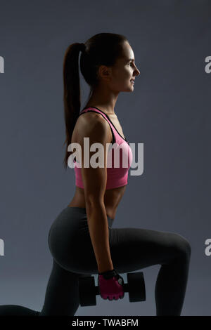 Seitenansicht des athletischen jungen brunette Mädchen im Sport Kleidung tun Hocke Übungen mit Hanteln für Gesäß auf grauem Hintergrund. Gerne hübsche Mädchen, das Training im Alltag fit und gesund zu bleiben Stockfoto