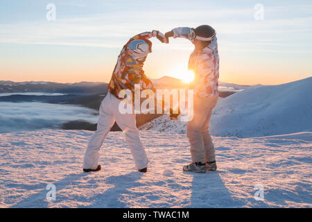 Paar schöne Hände halten in Form von Herzen auf schneebedeckten Gipfel bei Sonnenuntergang. Winterurlaub Stockfoto