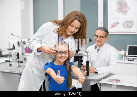 Funny Girl Wahl neue Brille in optischen Shop. Fröhliches Mädchen mit Kamera und zeigen anmelden Ok, während die weiblichen Optiker Brillen für besseren Anblick in der Klinik. Konzept der Korrektur des Sehvermögens. Stockfoto