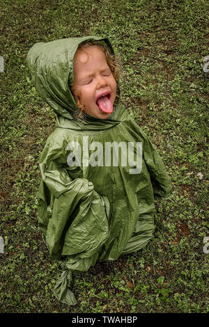 Wenig kleinkind Mädchen, Spielen im Regen, die versucht, es zu probieren, mit geschlossenen Augen und die Zunge heraus, mit grünen Regenjacke, auf grünem Gras Stockfoto