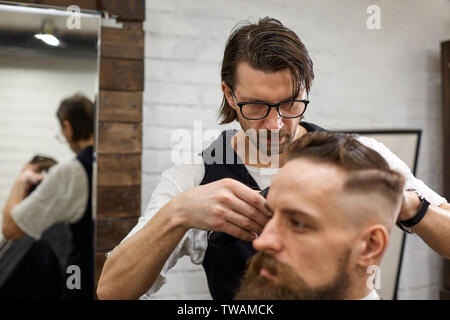 Meister schneidet Haare und Bart von Männern im Frisörsalon, macht Friseur Frisur für einen jungen Mann Stockfoto