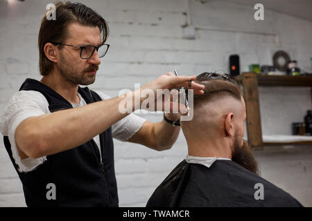 Meister schneidet Haare und Bart von Männern im Frisörsalon, macht Friseur Frisur für einen jungen Mann Stockfoto