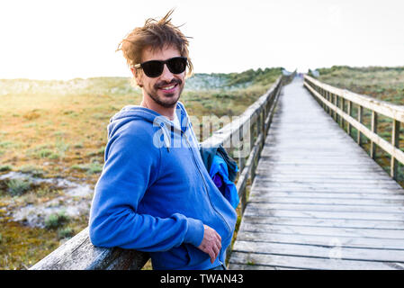 Hipster Spanisch aussehender junger Mann stand auf Sand Strand. Jungen attraktiven Mann steht auf hölzerne Brücke über Sanddünen auf Atlantischen Sandstrand in Stockfoto