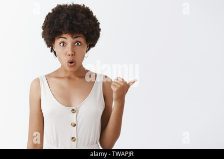 Studio shot beeindruckt und neugierig gewöhnlichen dunkelhäutige Freundin mit Afro Frisur in modischen weißen Overalls nach rechts mit Daumen, falzen Stockfoto