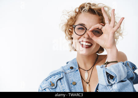 Nahaufnahme der charmante Sorglos charismatische junge Mädchen mit durchbohrten Nase in Gläsern und Lippenstift, okay Geste auf Auge und spähen durch ok Stockfoto