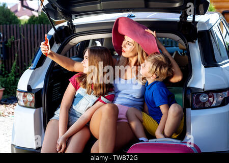 Mutter, Mädchen und Jungen laden bunte Koffer in den Kofferraum des Autos. Mom macht selfie auf einem Handy mit Ihren Kindern und bereit für die Reise. Stockfoto