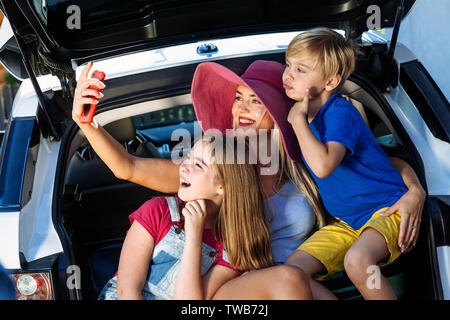 Mutter, Mädchen und Jungen laden bunte Koffer in den Kofferraum des Autos. Mom macht selfie auf einem Handy mit Ihren Kindern und bereit für die Reise. Stockfoto