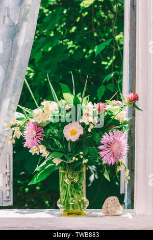 Ein Blumenstrauß von Clover, Kornblumen und Jasmin in einem Glas Vase und ein Herz aus Stein auf der Fensterbank eines offenen Fensters eines Dorfes Haus Stockfoto