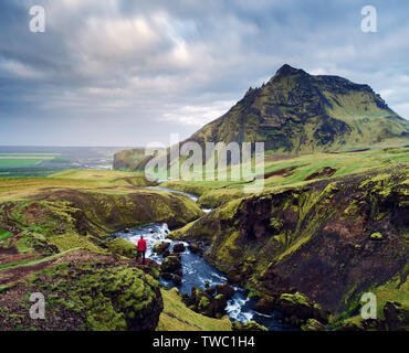 Skoga Fluss über den Wasserfall Skogafoss, Island, Europa. Reisende in einer roten Jacke steht auf einem Felsen und blickt auf die Berge Stockfoto