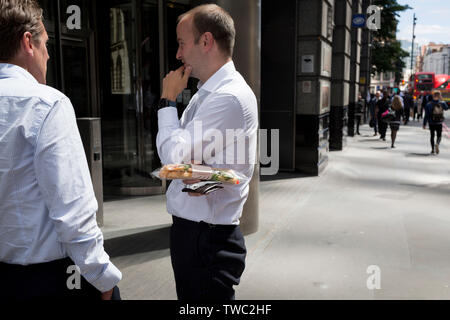 Ein Geschäftsmann hat einen Eingepackt takeaway Baguette, während im Gespräch mit einem Kollegen in die Stadt London, der Bezirk der Hauptstadt, die am 17. Juni 2019 in London, England. Stockfoto