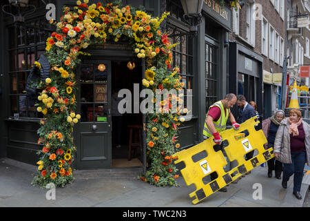 Neben einem Blumenarrangement über den Eingang des Engel & Crown Pub, Delivery Man kümmert sich um die Lieferung von neuen Aktien in den Keller an der St. Martin's Lane, die am 15. Juni 2019 in London, England. Stockfoto