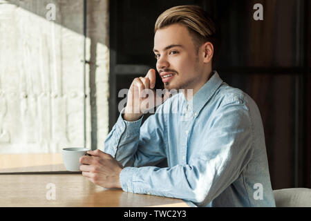 Seitenansicht Portrait von Happy hübscher junger bärtiger Mann in Jeans blau Shirt im Cafe sitzen, Kaffee trinken, haben arest und Telefonieren mit Stockfoto