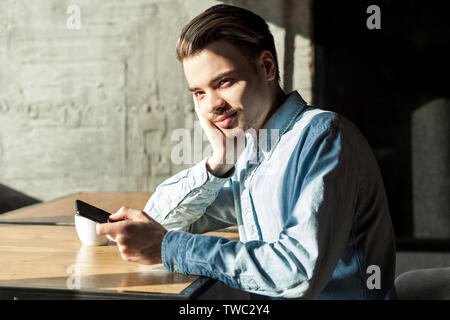 Seitenansicht Portrait von zufriedenen aufmerksamen jungen bärtigen Mann in Blau Jeanshemd im Cafe sitzen und halten Telefon und Denken. Ausruhen nach der Arbeit Stockfoto