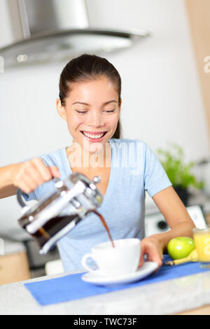 Frau essen Frühstück und trinken Kaffee am Morgen. Mädchen gießt Kaffee aus der französischen Presse zu Hause in der Küche. Schönen gemischten Rennen asiatischen Kaukasischen weibliche Modell lächelte glücklich. Stockfoto