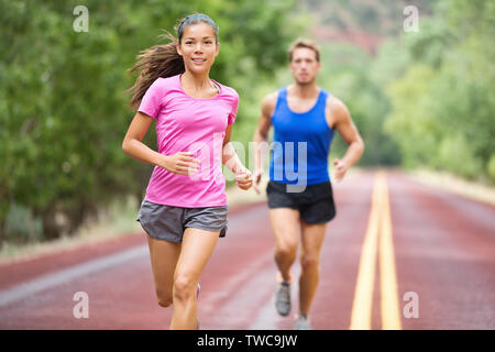 Ausführen von jungen gemischten Rennen paar Training für Marathon draußen auf der Straße. Glücklich lächelnde schönen gemischten Rennen weibliche model in der Stattlichen kaukasischen männliches Modell im Hintergrund verschwommen, auf denen fokussierte Stockfoto