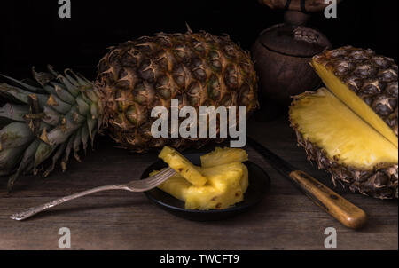 In Scheiben geschnittene Ananas auf alten rustikalen Schreibtisch mit Messer und mit ganzen Ananas hinter sich. Schwarzen Hintergrund. Stockfoto